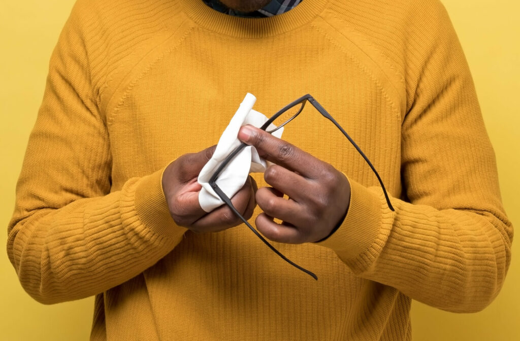 A man in a yellow sweater cleaning his glasses using a white microfiber cloth against a yellow background.