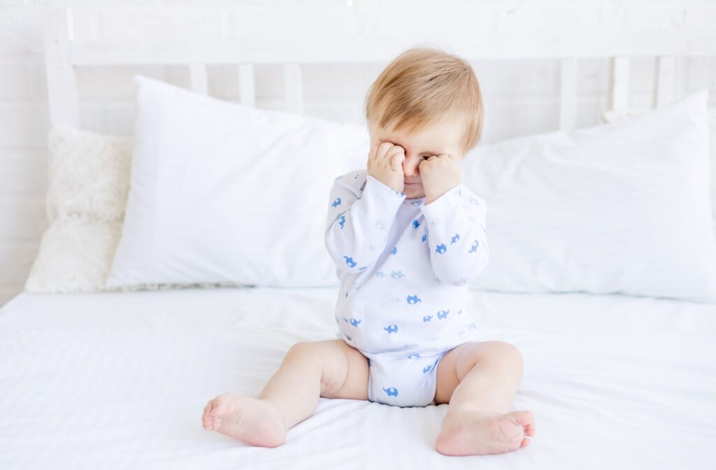 A baby in white pyjamas rubbing their eyes with both hands.