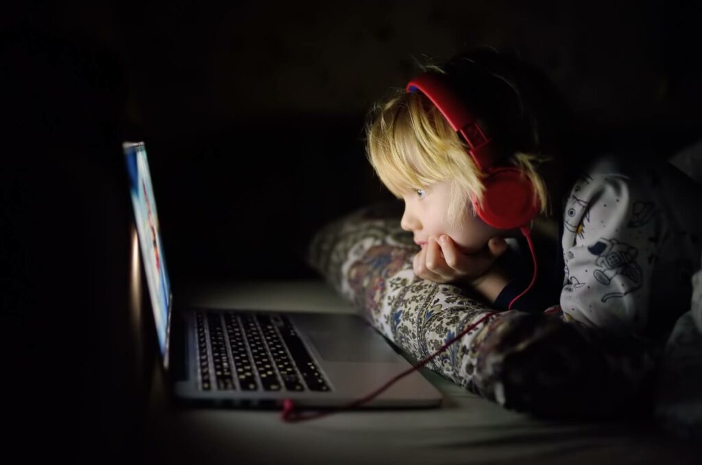 A child sitting in the dark, wearing headphones and watching something on a laptop.