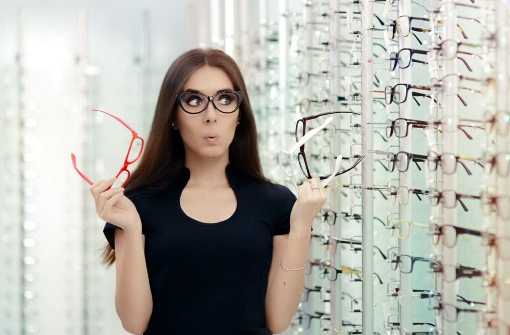 A person playfully trying on glasses, holding two frames in their hands while standing in front of a wall of eyeglasses.