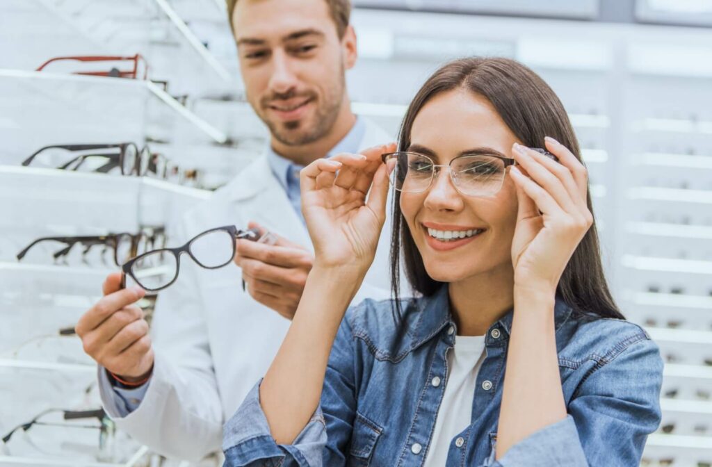 A smiling person trying on new glasses with the help of an optician in an eyewear store.