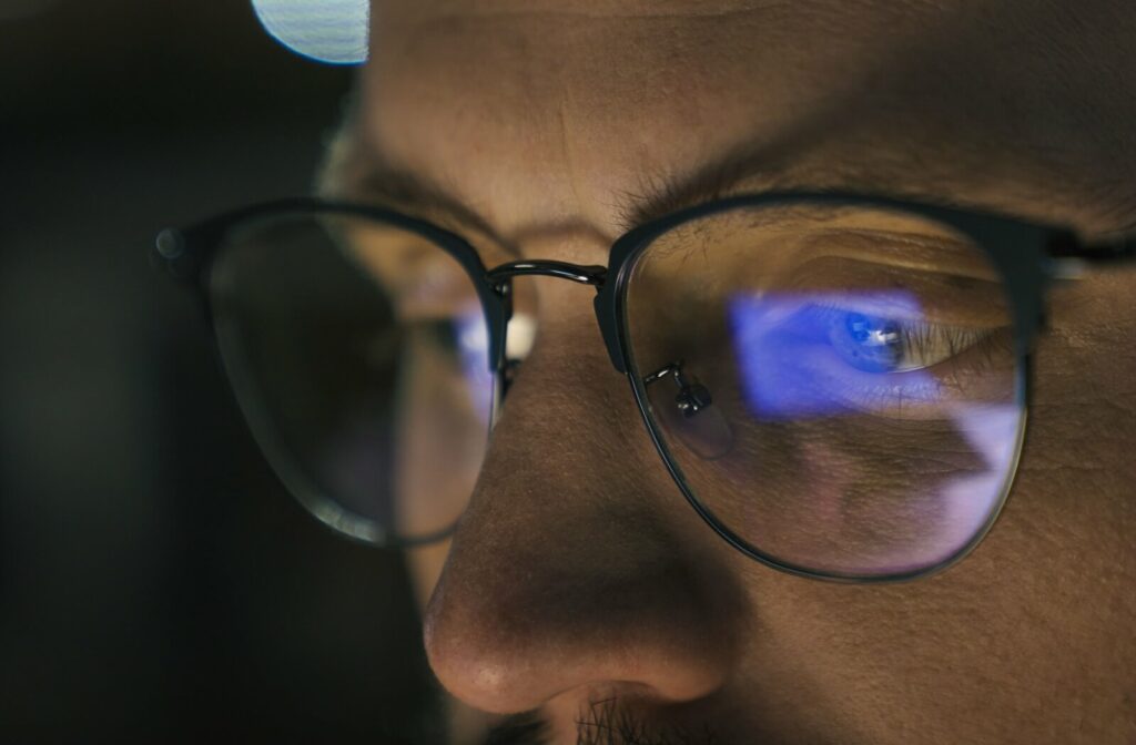 Close-up of a person wearing glasses with blue light from a digital screen reflecting on the lenses while looking at a monitor.