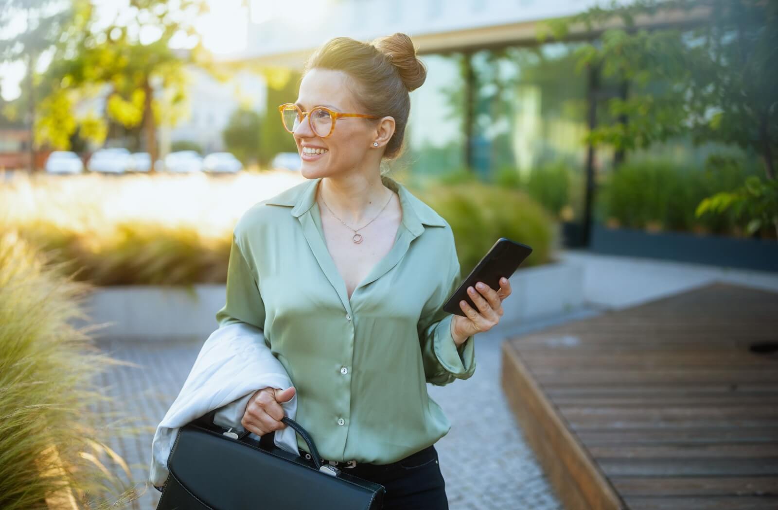 A person wearing round, tortoiseshell glasses and a green blouse smiling while walking outdoors and holding a smartphone and a briefcase.